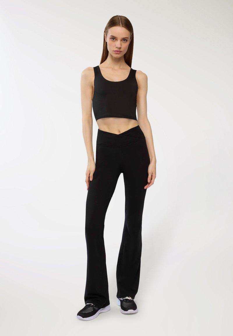 Top corto nero e pantaloni a vita alta svasati neri, entrambi realizzati in tessuto morbido. Modello che indossa scarpe sportive nere con dettagli in rete.