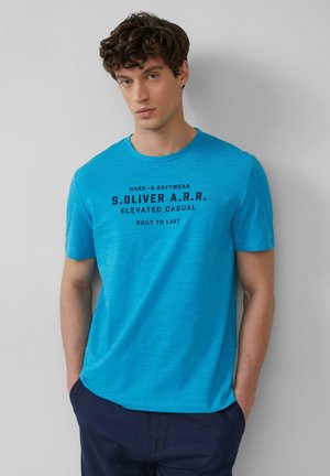 Turquoise crewneck t-shirt van lichtgewicht stof, met een ontwerp in zwarte tekst en korte mouwen. Gecombineerd met marineblauwe broeken.