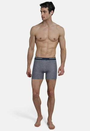 Muscular man standing barefoot wearing gray fitted boxer briefs with a black waistband labeled "bugatti," looking to his left.