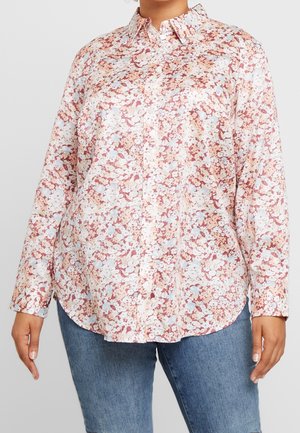 Mujer que lleva una camisa de manga larga con botones y un estampado floral en rosa, blanco y azul claro combinada con vaqueros azules.