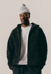 Man wearing gray knit hat, black zip-up jacket, light gray sweatshirt with letters, and dark pants, standing against a plain background.