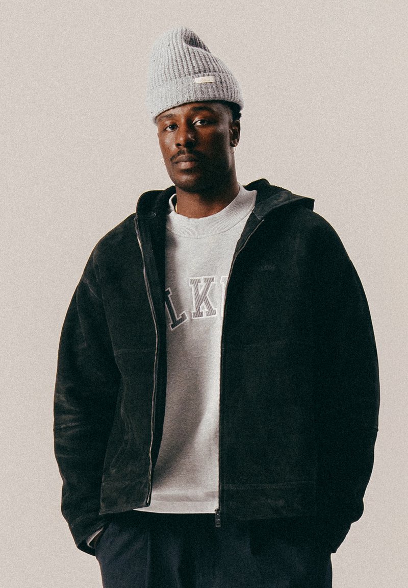 Man wearing gray knit hat, black zip-up jacket, light gray sweatshirt with letters, and dark pants, standing against a plain background.