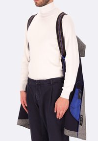 Cappotto navy con colletto di lana grigia, caratterizzato da foderatura blu e dettagli con cinturino. Indossato sopra un dolcevita bianco e pantaloni scuri.