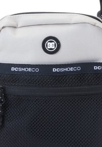 Zaino a rete nero con una sezione superiore grigia chiara, logo DC e cerniere. Il materiale sembra resistente con elementi di design strutturati.