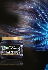Il barattolo Garnier Ultra Dolce Hair Remedy presenta un contenitore nero con un coperchio dorato, etichettato con grafiche floreali. Sullo sfondo ci sono ciocche di capelli fluenti con riflessi blu.