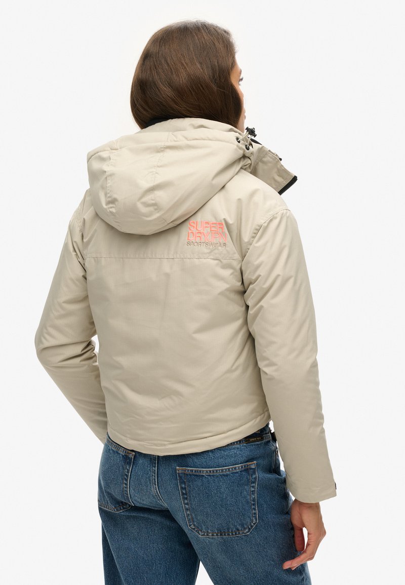 Beżowa kurtka puchowa z kapturem, z czarnym zamkiem błyskawicznym i pomarańczowym logo "Superdry Sportswear" na plecach, w zestawie z niebieskimi dżinsami.