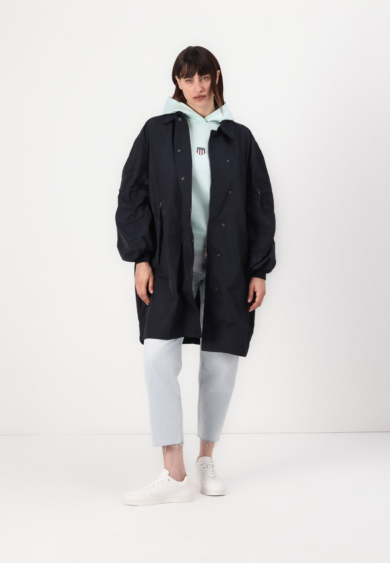 Chaqueta extragrande en color navy con cuello puntiagudo, cierre de botones y grandes bolsillos, superpuesta a una sudadera ligera en color menta y vaqueros azul claro. Zapatillas blancas.