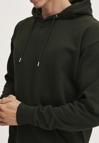 Felpa verde scuro con cappuccio a coulisse e punte metalliche, polsini a costine e una texture morbida. La vestibilità è rilassata e informale.