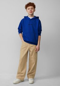 Blauwe hoodie met een zwart logo, gecombineerd met beige wijde broek en witte sneakers, tegen een neutrale achtergrond.