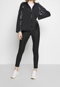 Svart, lättvikts vindjacka med dragkedja, huva och vita textaccenter på ärmarna. Kombineras med svarta leggings och vita sneakers.