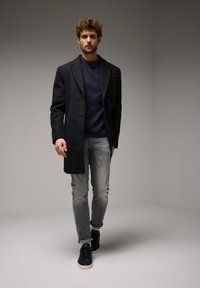 Šedý vlněný kabát s klopovým límcem, doplněný tmavě modrým svetr, ošuntělými světlými džíny a černými teniskami. Pozadí je minimalistické.