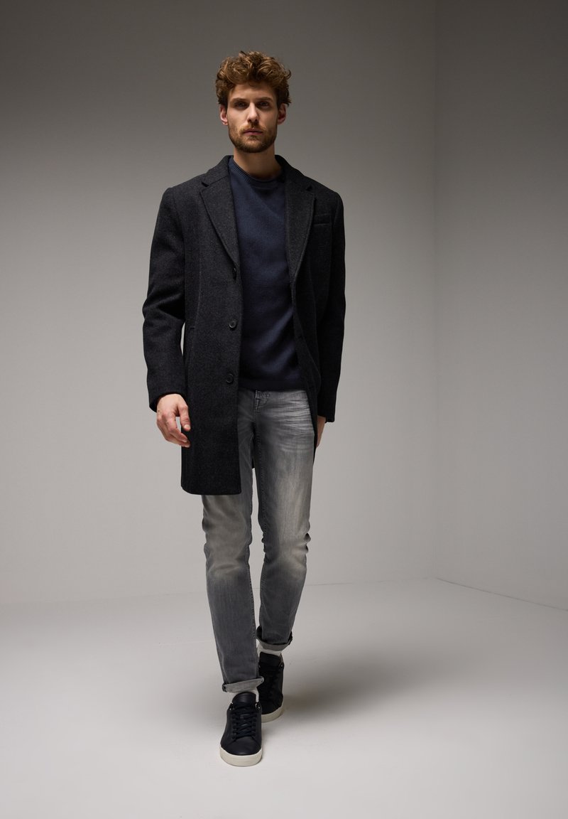 Šedý vlněný kabát s klopovým límcem, doplněný tmavě modrým svetr, ošuntělými světlými džíny a černými teniskami. Pozadí je minimalistické.