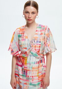 Robe chemise multicolore avec des manches courtes à volants, présentant un motif mixte de rayures et de carrés, fabriquée à partir d'un tissu léger et transparent.