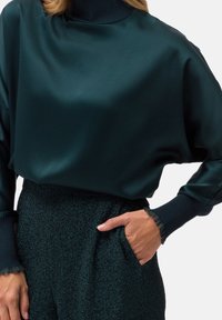 Top de manga larga en verde azulado oscuro con cuello alto, hecho de tela suave. Presenta puños acanalados con bordes con volantes y una silueta relajada.