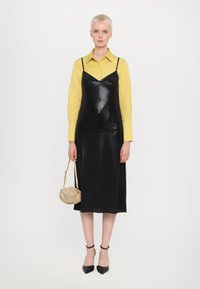 Schwarzes, glänzendes Kleid mit dünnen Trägern über einem leuchtend gelben Shirt. Das Model hält eine kleine, beige verzierten Tasche und trägt schwarze High Heels.