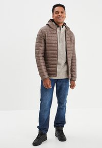 Braune isolierte Jacke mit horizontalem Steppmuster, grauer Pullover, blaue Jeans und schwarze High-Top-Sneaker.