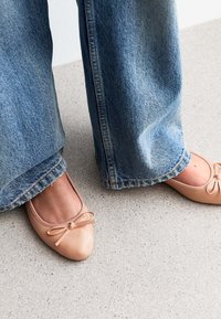 Beige patent leather ballet flats with a small bow detail, worn with wide-leg blue denim jeans, on a light concrete surface.