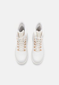 Et par hvide vinterstøvler med gyldne snørehuller, hvide snørebånd, tykke såler og blød fleece-foring, vist oppefra på en ensfarvet baggrund.