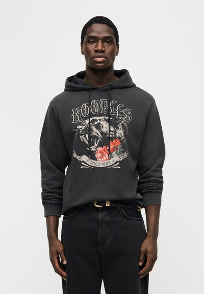 Jeune homme portant un sweat à capuche gris foncé avec une panthère rugissante, des fleurs rouges et le texte « Kooples Wild Tour », associé à un jean noir et une ceinture.