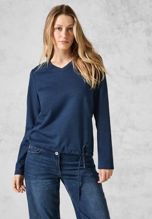 Blå långärmad topp i texturerad tyg med V-ringning, insnörd nederkant och justerbar dragsko; kombinerad med mörka jeans.