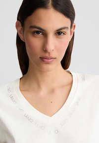 T-shirt blanc à col en V avec une texture en coton lisse, arborant un motif de logo métallique autour de l'encolure. Design simple et moderne.