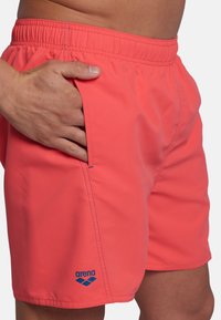Pantaloni da bagno corallo realizzati in materiale asciugatura rapida; dotati di tasca laterale, vita elastica e accentuazione blu del logo sul lato sinistro inferiore.