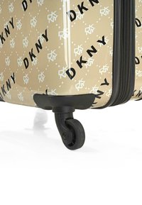 Maleta rígida en beige con estampado del logo de DKNY en negro y blanco. Cuenta con ruedas negras duraderas y cierre de cremallera para un fácil transporte.