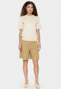 Beige polo tričko s krátkym rukávom, golierom a tromi gombíkmi, v kombinácii s voľnými khaki šortkami a svetlobežovými plochými topánkami.