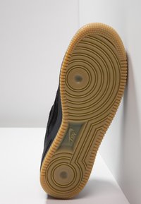 Sola di una sneaker nera Nike con suola in gomma color cuoio, caratterizzata da cerchi concentrici e logo Nike, posizionata verticalmente su una superficie bianca.