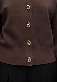 Rudas megztas kardiganas su V formos iškirpte ir šešiais blizgiais sidabriniais mygtukais, turintis abstrakčią dizainą. Apačia šiek tiek rifoja.