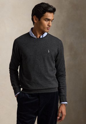 Mand iført mørkegrå crew neck sweater over lyseblå skjorte med krave, stående med den ene hånd i lommen, kiggende mod højre.