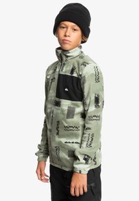 Grüner Fleece-Pullover mit schwarzem Brustpatch, der abstrakte schwarze Muster aufweist. Hoher Kragen und lange Ärmel, getragen mit einer schwarzen Mütze.