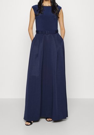 Kobieta ubrana w bezrękawnikową, granatową suknię sięgającą podłogi z paskiem, stojąca z rękami w kieszeniach, widoczne czarne sandały na otwartym palcu.