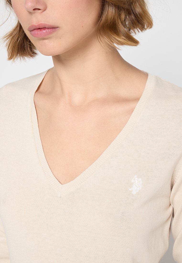 Beige V-hals trui van zachte stof met een fitted design; heeft een klein wit logo op de linkerkant van de borst en ribgebreide manchetten.