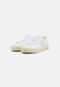 Hvite lerretsSneakers med beige gummisåler, med en fremtredende beige logo på siden, og standard snøresystem med lufthull.