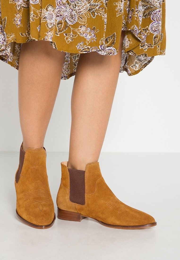 Femme portant des bottines en suède couleur moutarde avec des talons bas empilés et des panneaux latéraux élastiques, associées à une robe fleurie moutarde.