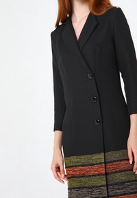 Svart kavajklänning med skräddarsydd silhuett, prydd med tre svarta knappar och en livlig randig vävd fåll i grön, röd och brun.