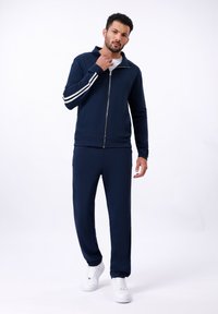 Survêtement marine comprenant une veste zippée avec des rayures blanches sur les manches et un pantalon assorti. Fabriqué en tissu doux, accompagné de baskets blanches.