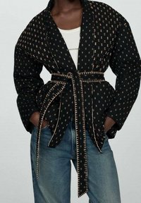 Czarna kurtka w wzory z złotymi akcentami, o kroju typu wrap, z szerokimi rękawami i paskiem w talii. Materiał wydaje się teksturowany i lekki.