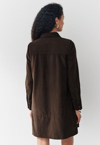 Brązowa sukienka koszulowa wykonana z tkaniny o fakturze, z kołnierzykiem, mankietami na guziki i długością do połowy łydki. Widok od tyłu.