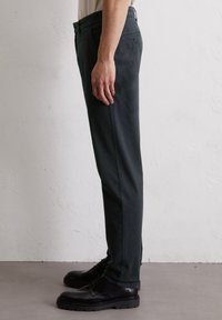 Pantalon slim vert foncé avec une texture lisse, doté de poches latérales et d'un détail en bouton, associé à des chaussures noires à lacets.