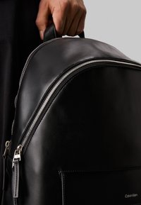 Zaino in pelle nera con forma arrotondata, caratterizzato da una zip argentata e una superficie liscia. Include un logo visibile e una maniglia.