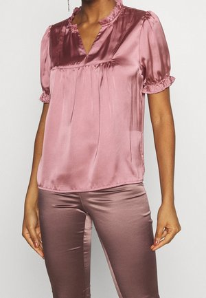 Kvinde iført en skinnende mauve kortærmet bluse med flæsekrave og matchende satinpants mod en ensfarvet baggrund.