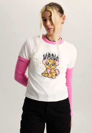Jonge vrouw met lang blond haar draagt een wit en roze gelaagde longsleeve met een cartoonhond met vlammen en glitters op de voorkant.