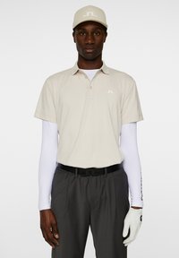 Krämfärgad polo med korta ärmar, logotyp på vänster bröst, kombinerad med mörkgrå byxor och en vit långärmad undertröja. Vita handskar.