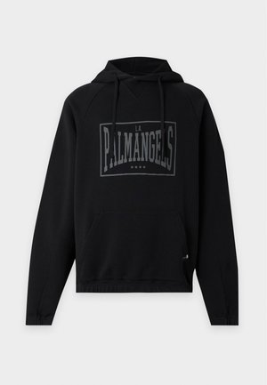 Homme portant un sweat à capuche noir avec texte, pantalon blanc à jambes larges avec boutons latéraux et chaussures noires, debout sur un fond uni.