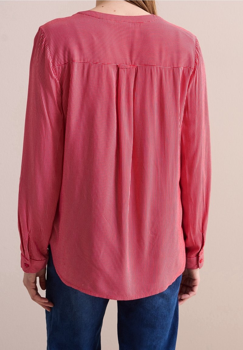 Red and white striped long-sleeve shirt with a round neckline, back pleat detail, and buttoned cuffs, paired with blue denim.