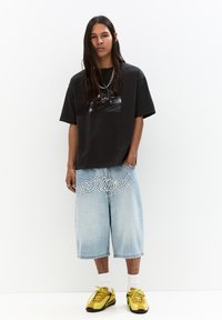 Black short-sleeve T-shirt with a graphic print; light blue baggy denim shorts with white embroidered text; yellow sneakers.