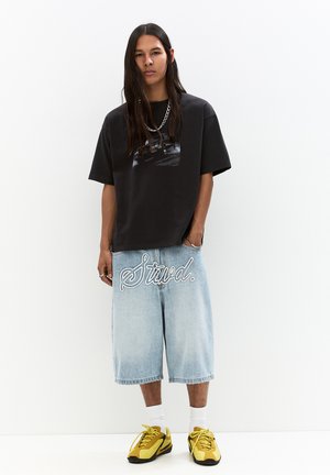Black short-sleeve T-shirt with a graphic print; light blue baggy denim shorts with white embroidered text; yellow sneakers.