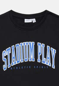 Felpa nera con collo rotondo, caratterizzata da un audace testo bianco e blu che recita "STADIUM PLAY" e un testo più piccolo sotto che dice "ATHLETIC SPIRIT."